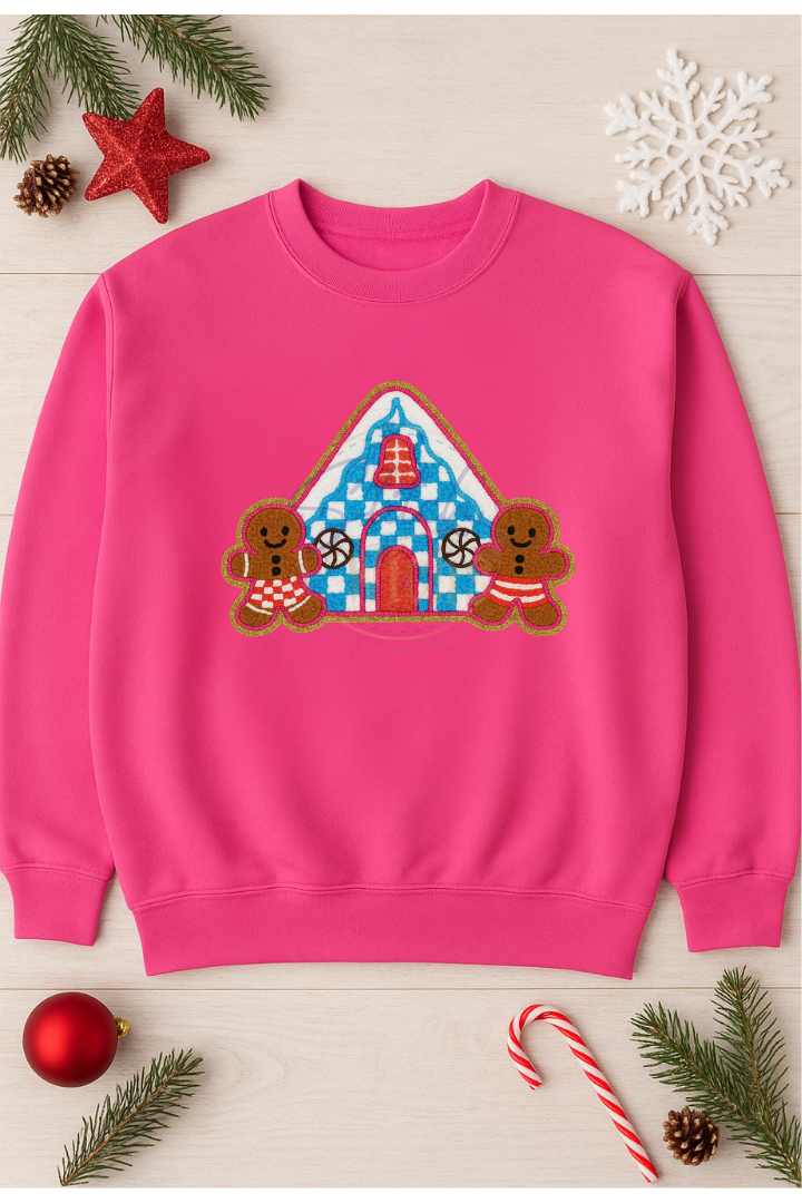 Pink sweatshirt with gingerbread house design surrounded by Christmas decorations on a light wooden surface.